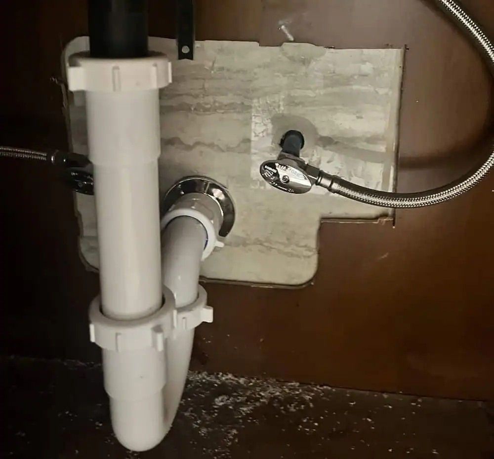 White PVC pipes and metal braided water lines under a sink with a hole cut in the wood paneling.