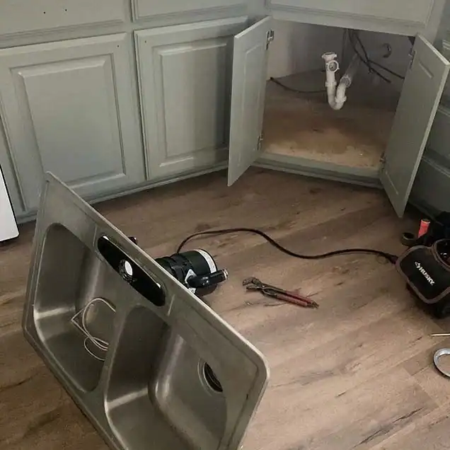 Double kitchen sink removed on floor with plumbing tools near open cabinet with exposed pipes underneath.