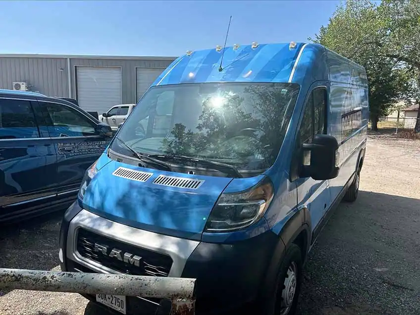 Blue Ram delivery van parked on gravel near a building and trees under a clear sky.