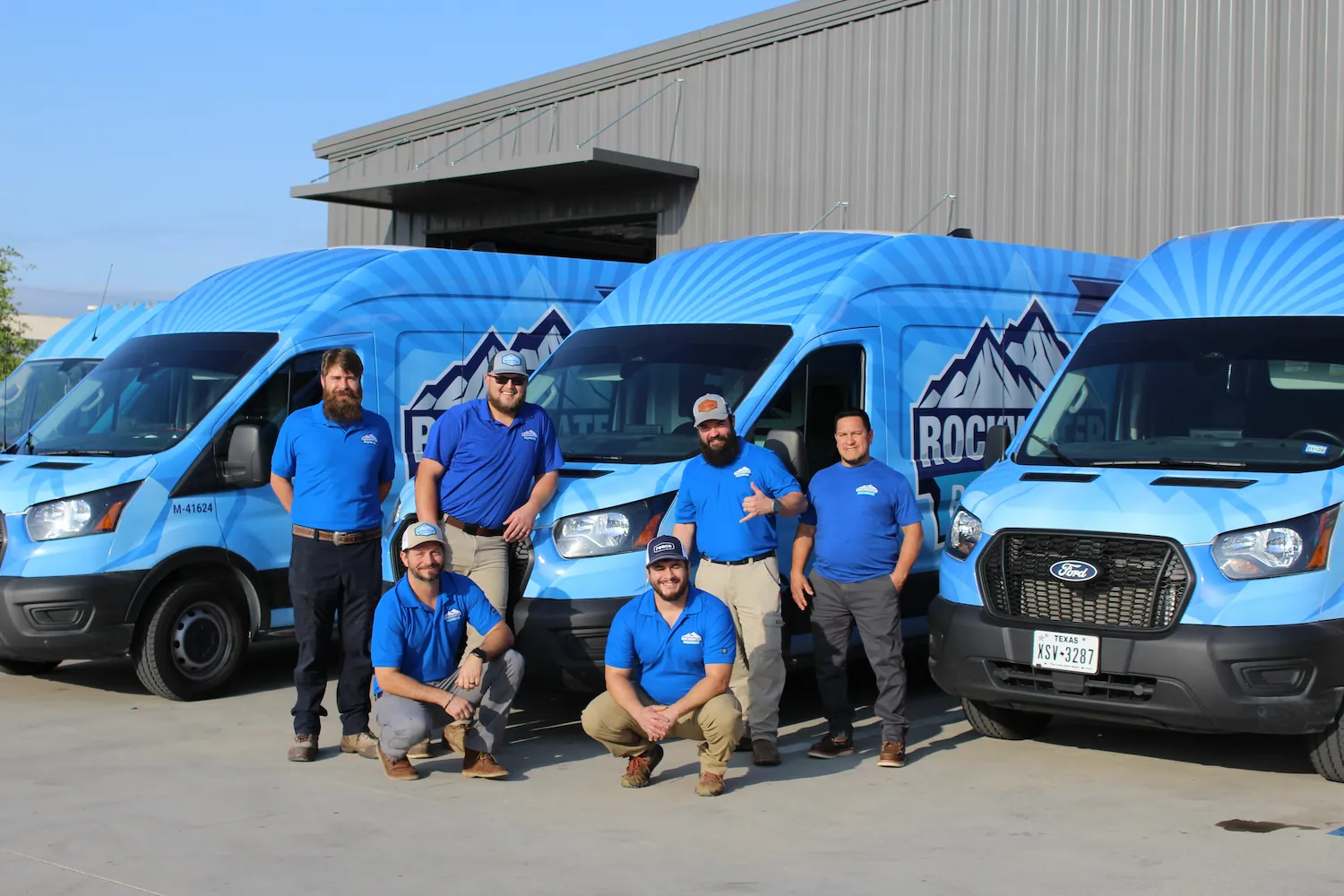Six men in blue shirts stand and kneel in front of three blue vans with Rocky Mountain logos outside a building.