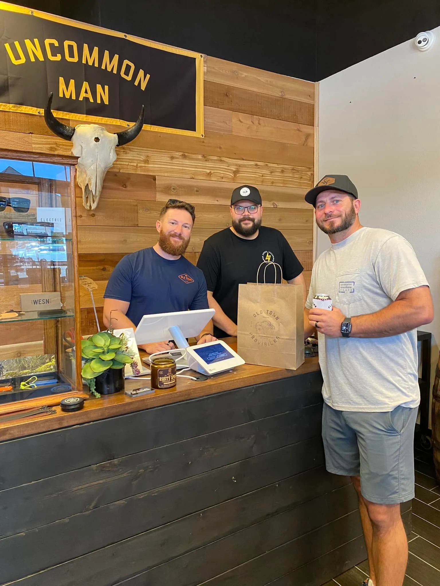 Three men smiling inside a rustic store with wood walls and an 'Uncommon Man' sign above them.