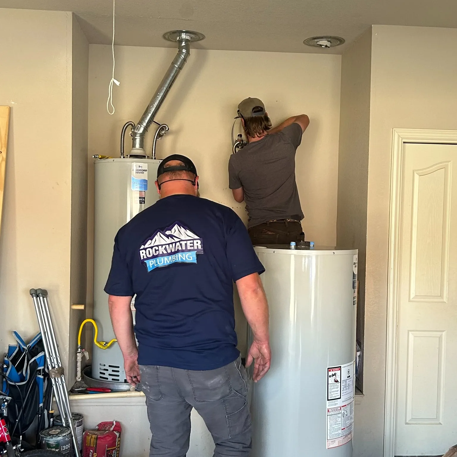 Two plumbers working on water heaters in a utility closet, one wearing a Rockwater Plumbing shirt.