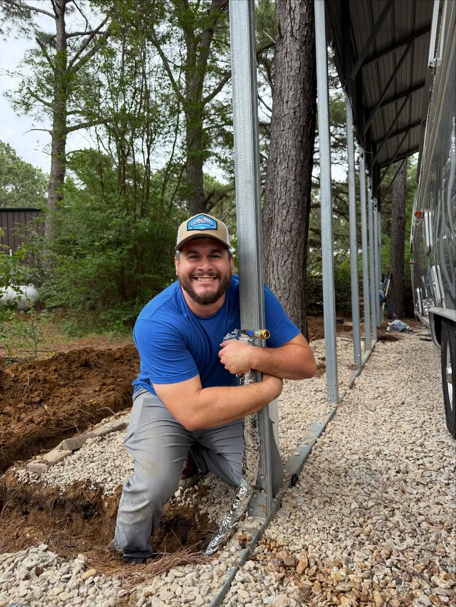 Smiling man in blue shirt and cap kneeling on gravel beside a metal post under a metal roof.