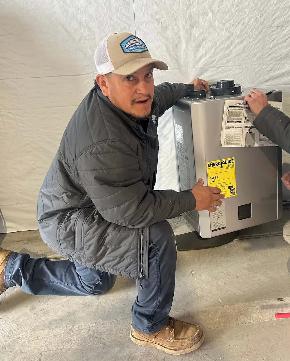 Man wearing a Rockwater Plumbing cap kneels beside a machine with an EnergyGuide label.