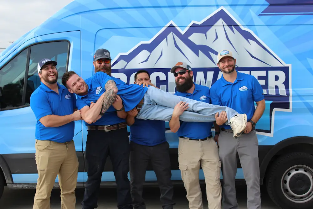Five men in blue shirts hold up a smiling man in jeans in front of a blue van with mountain logo.