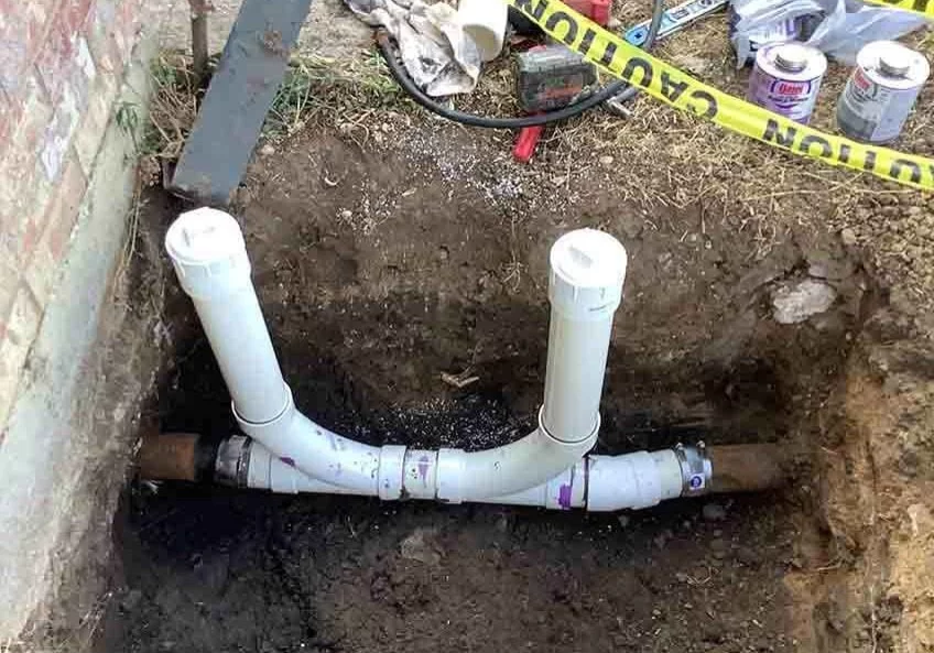 White PVC piping installed in a dug trench next to a brick wall with caution tape nearby.