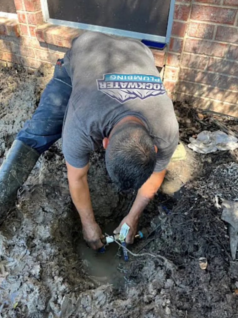 Plumber in gray shirt working with pipes in muddy ground outside a brick building.