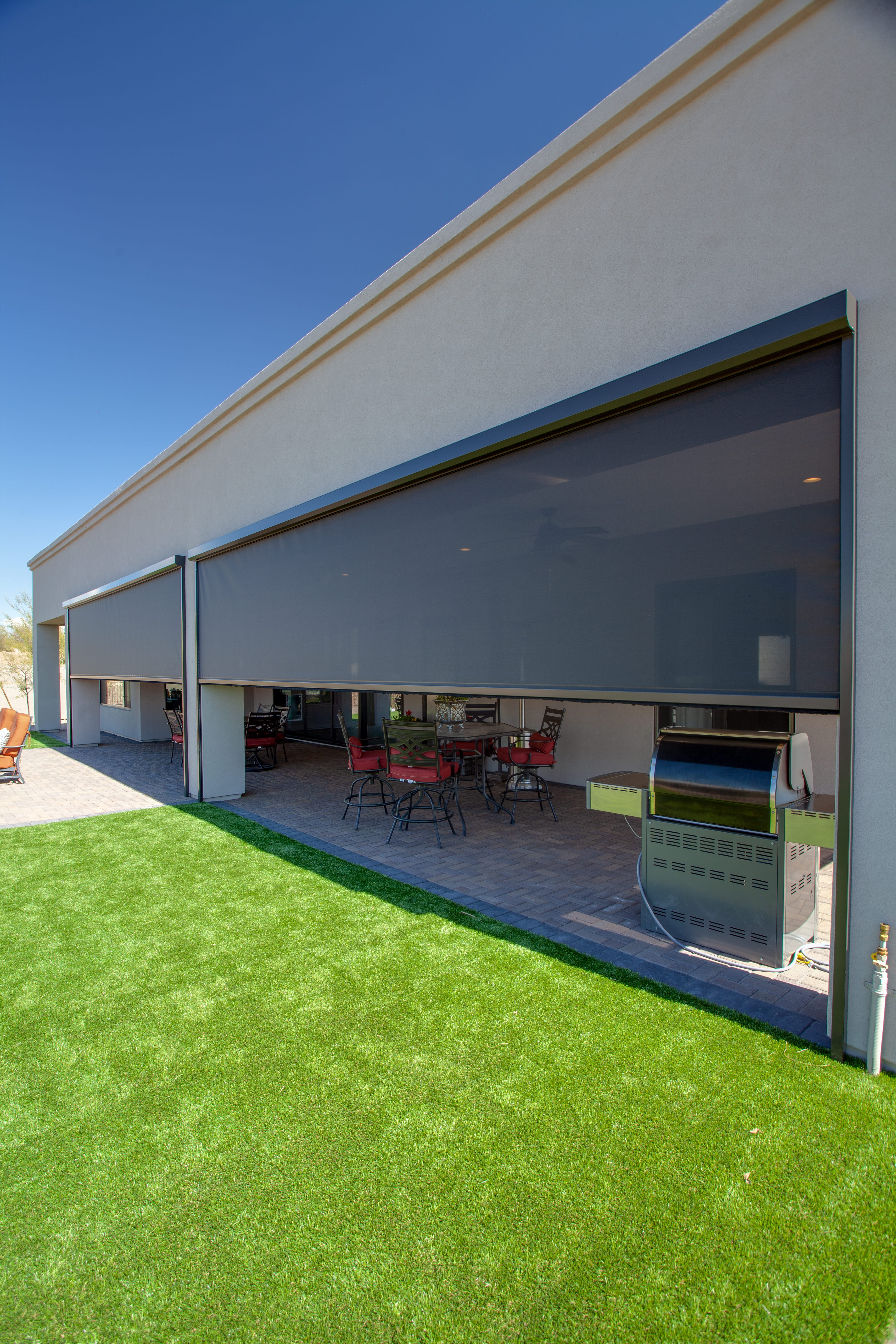 Exterior solar screen shades lowered over a covered patio with a dining set and grill