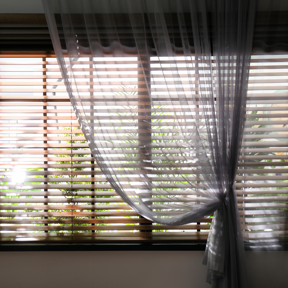 Wood blinds behind a sheer gray curtain on a sunny window