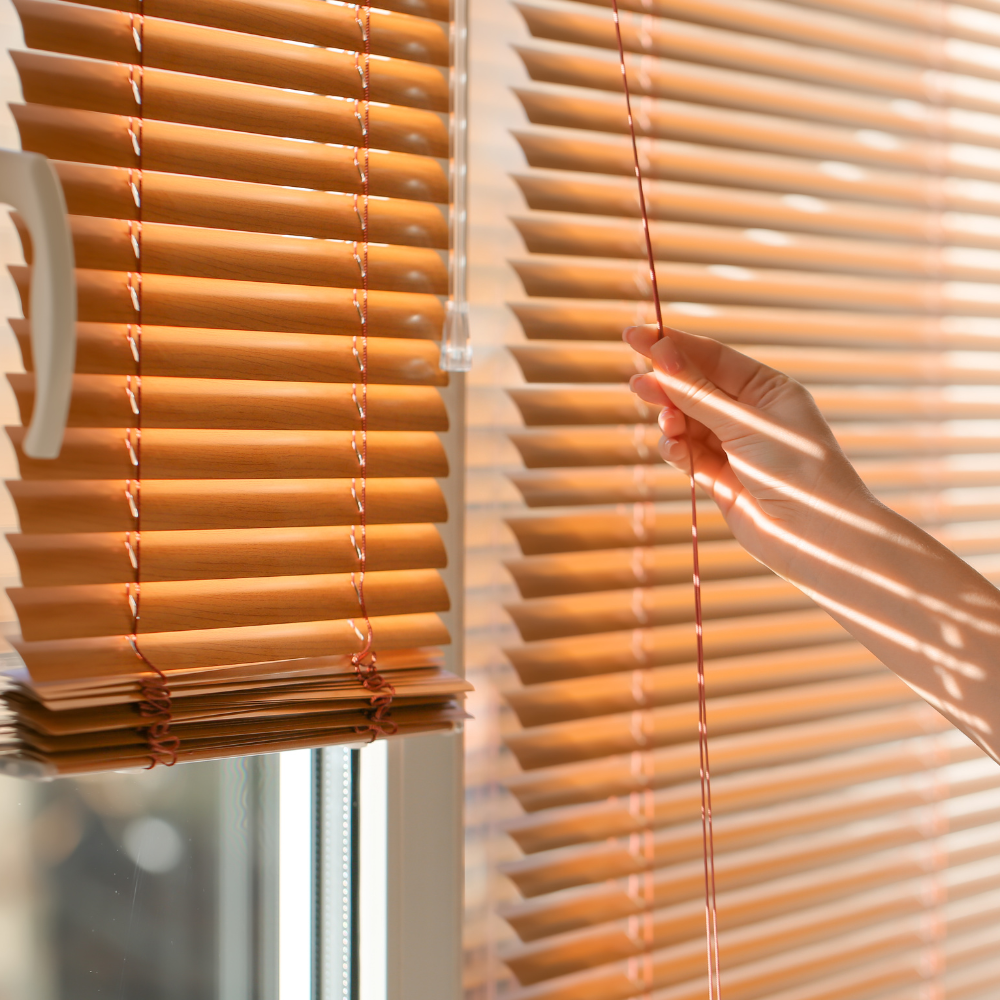 Hand pulling the cord on tan composite blinds lit by warm sunlight