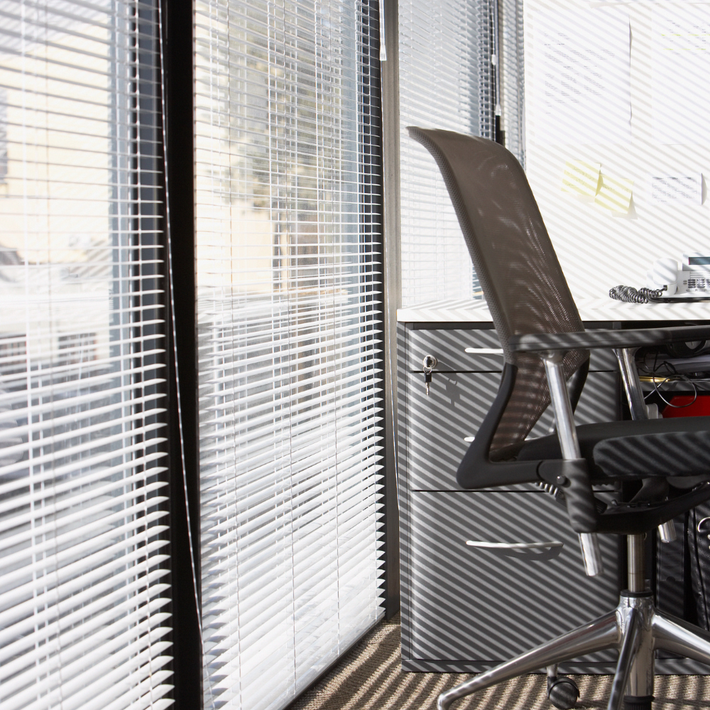 White vinyl mini blinds on sunny office windows beside a mesh desk chair