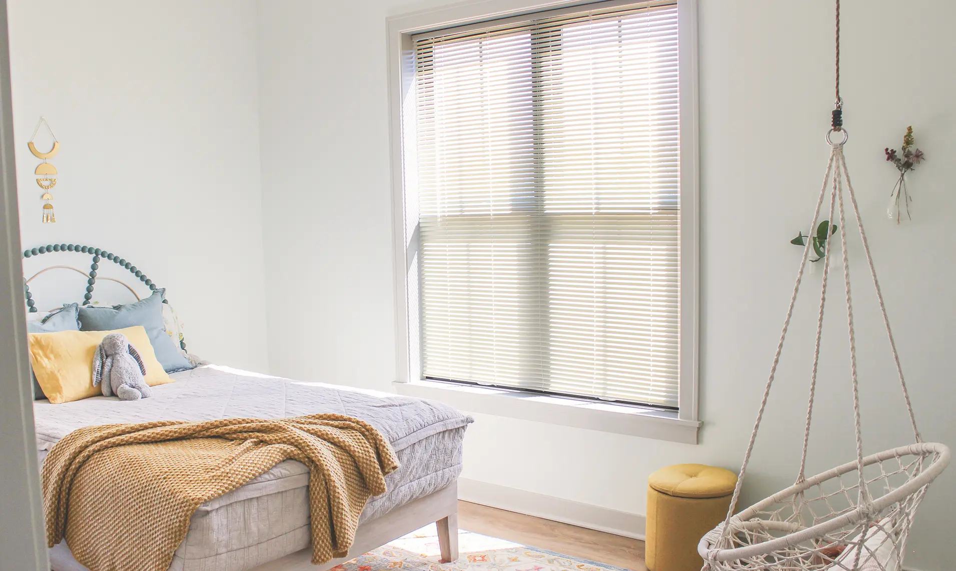 White vinyl horizontal blinds on a bright bedroom window beside a bed and hanging chair