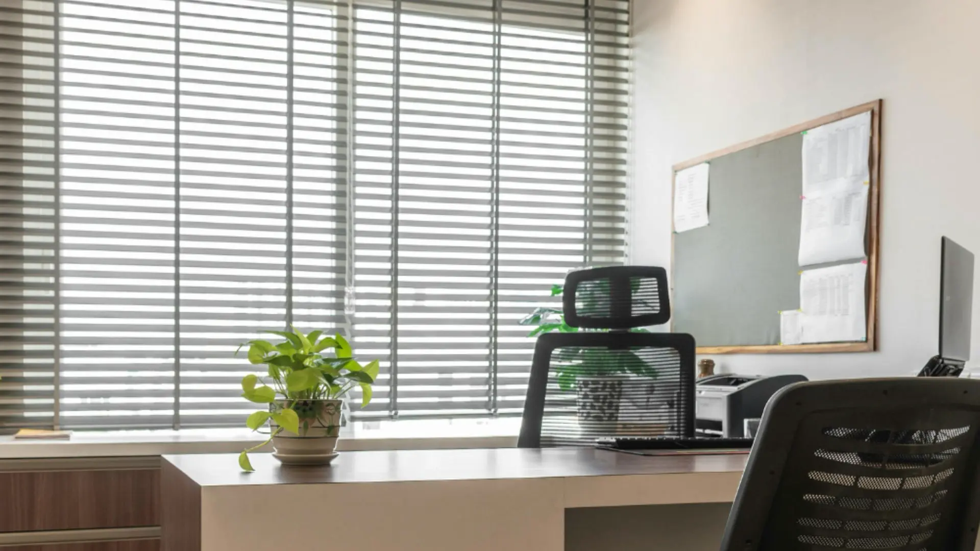 Gray horizontal blinds on a large window in a modern office with desk and mesh chairs