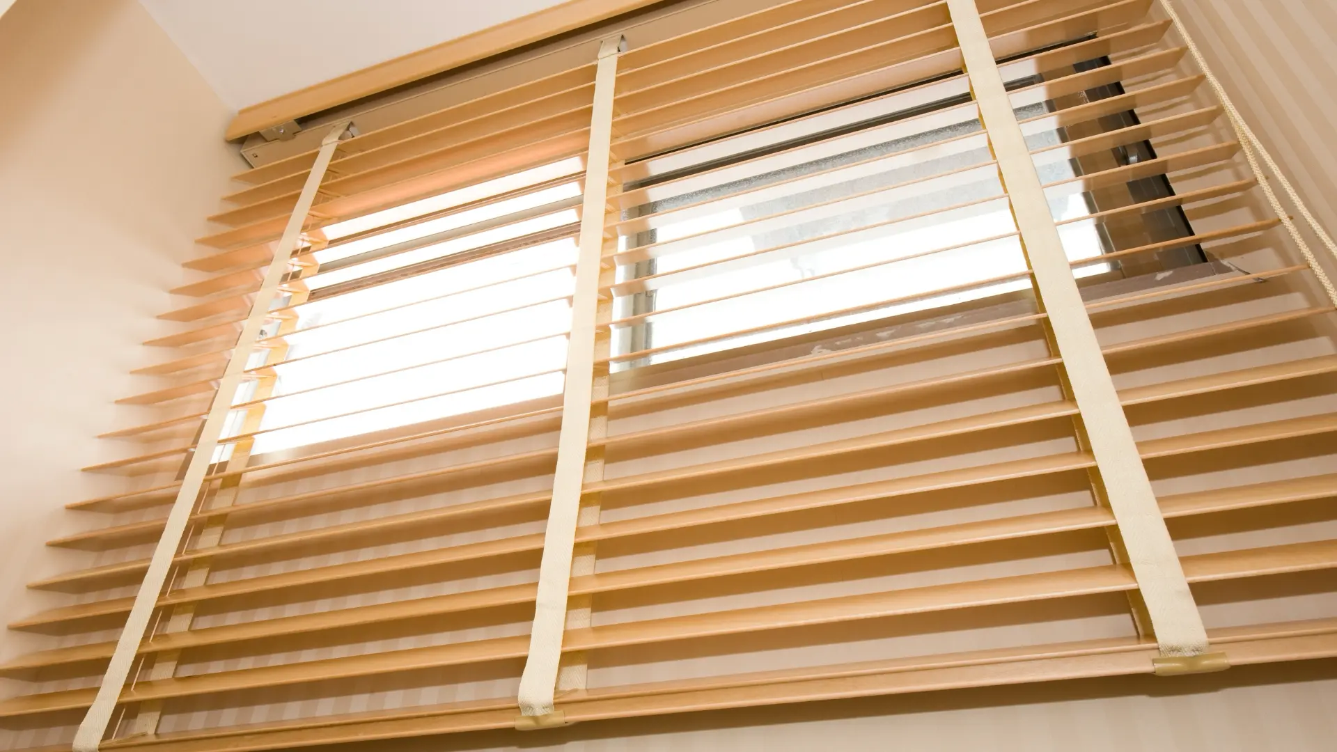 Natural wood blinds with cloth ladder tapes covering a window, viewed from below