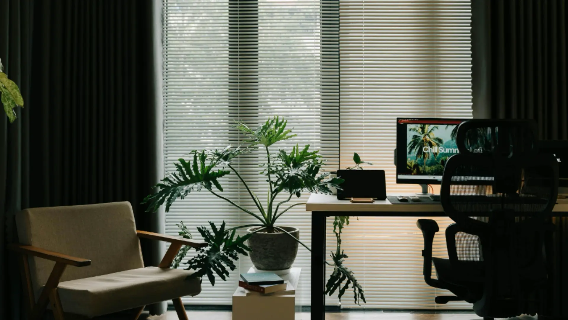 Home office with aluminum mini blinds behind a desk, potted plant and lounge chair