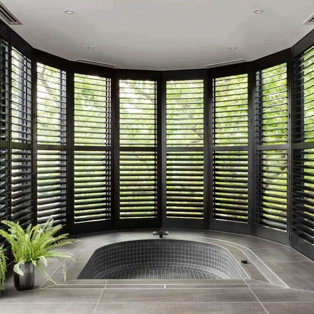 Black plantation shutters lining a curved bay window around a sunken bathtub