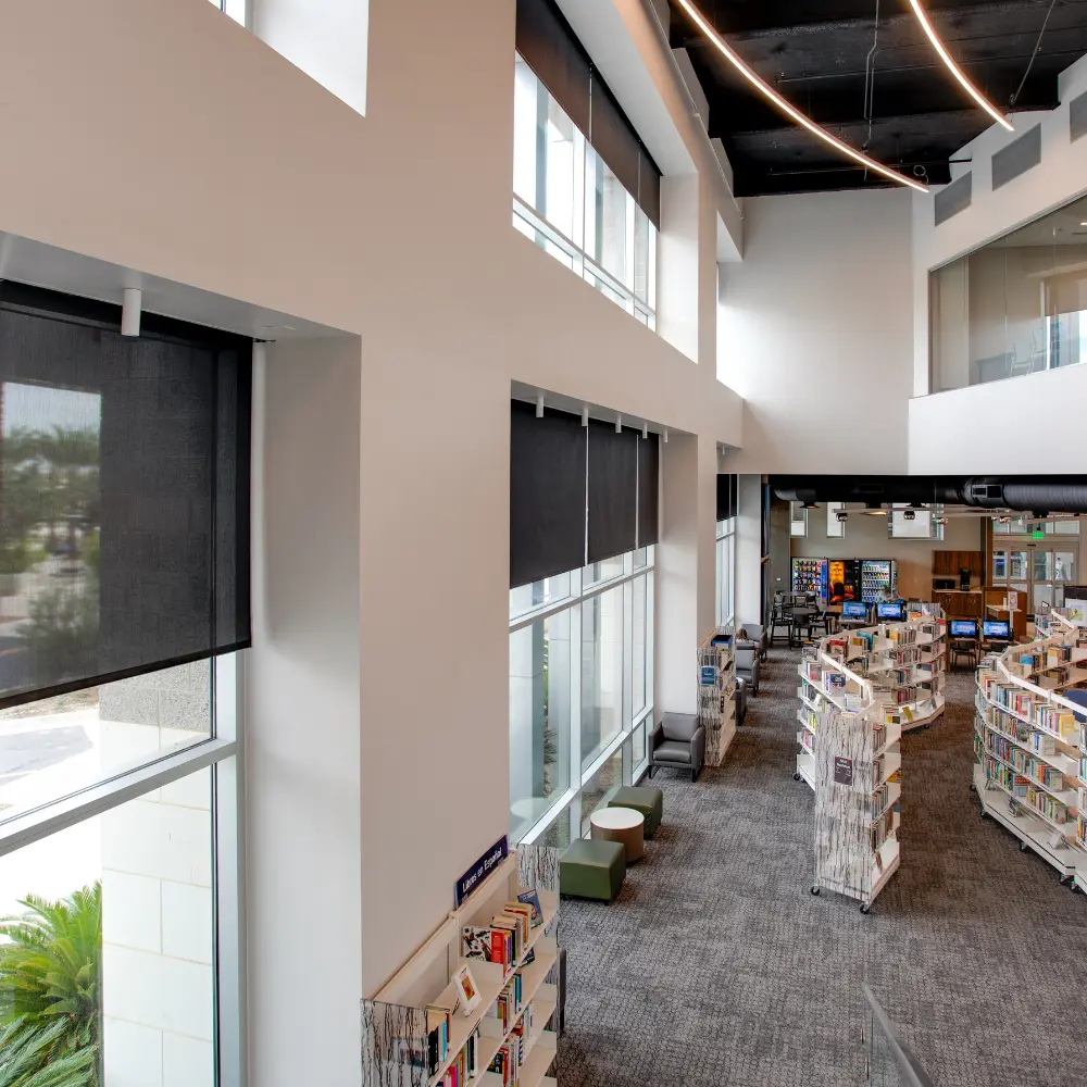 Dark solar shades on tall windows in a two-story school library with bookshelves