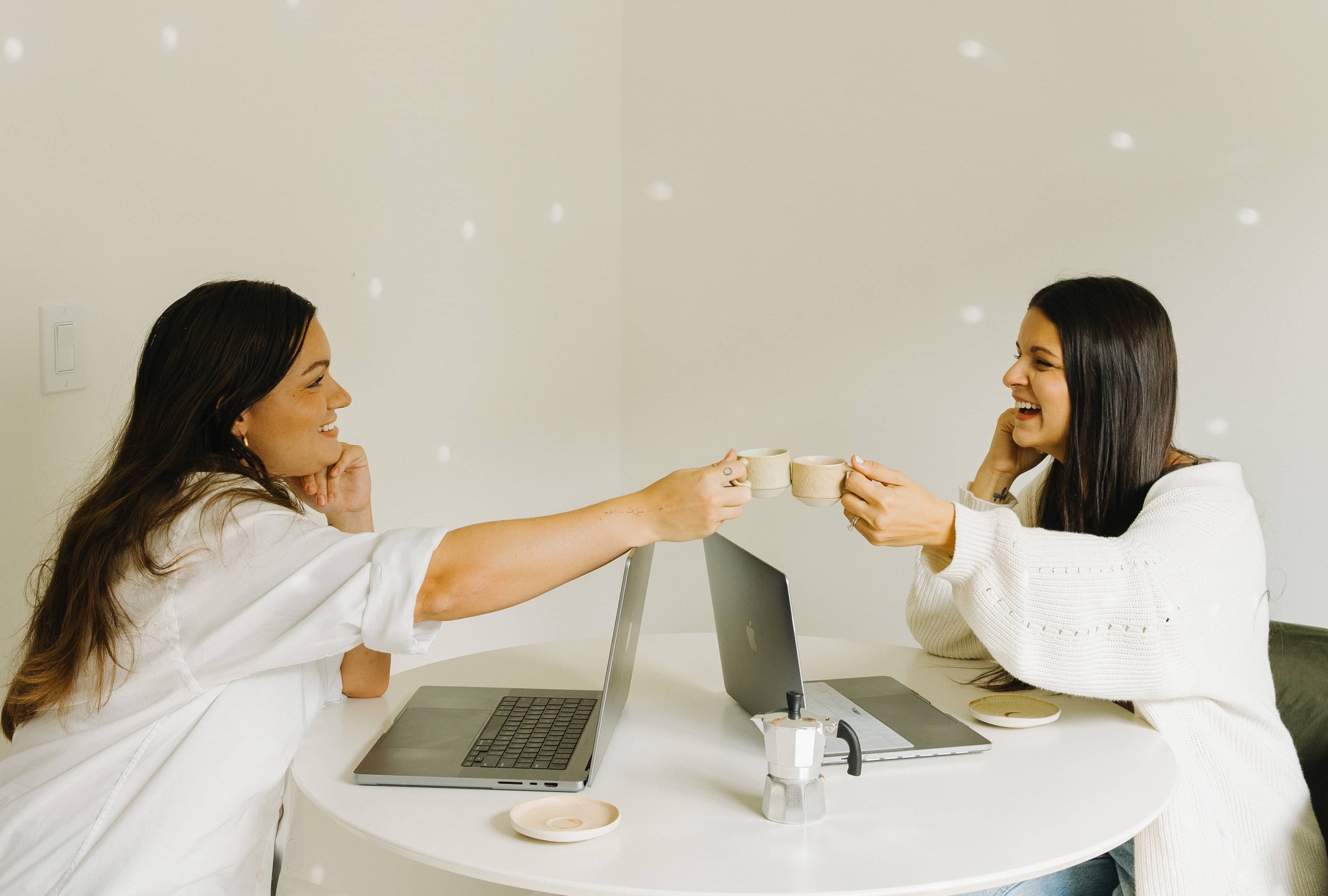 Two women toasting espresso cups over their laptops