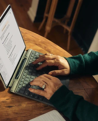 Person typing on a tablet keyboard at a wooden round table with a notebook nearby.
