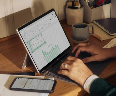 Person typing on tablet keyboard showing spreadsheet with charts on a wooden desk with phone and mug nearby.