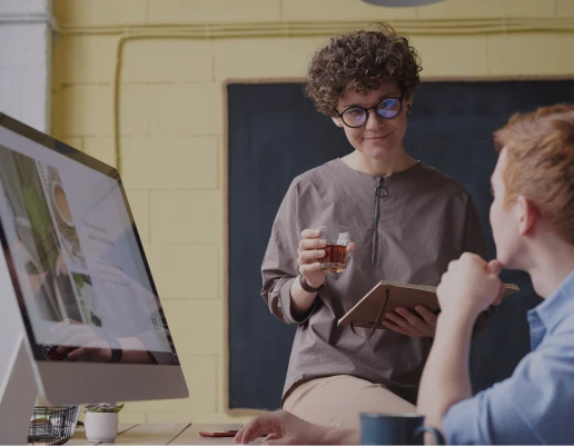 Woman holding drink and notebook talks to man sitting by computer in casual office space.