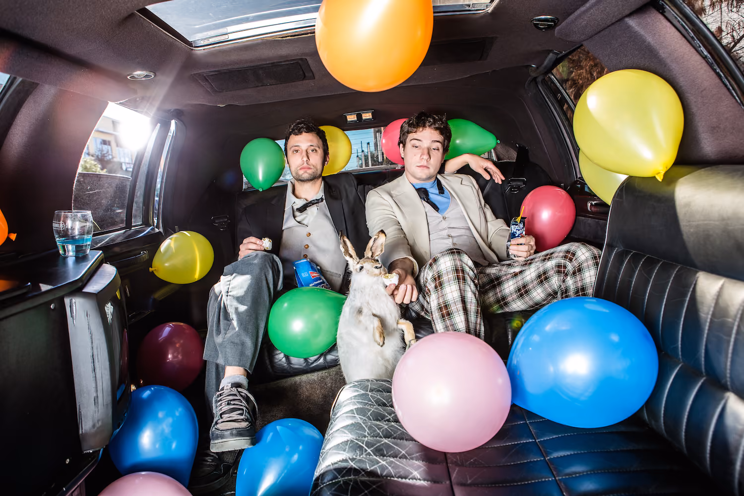 Two men in suits posing in a limousine filled with balloons and a small dog