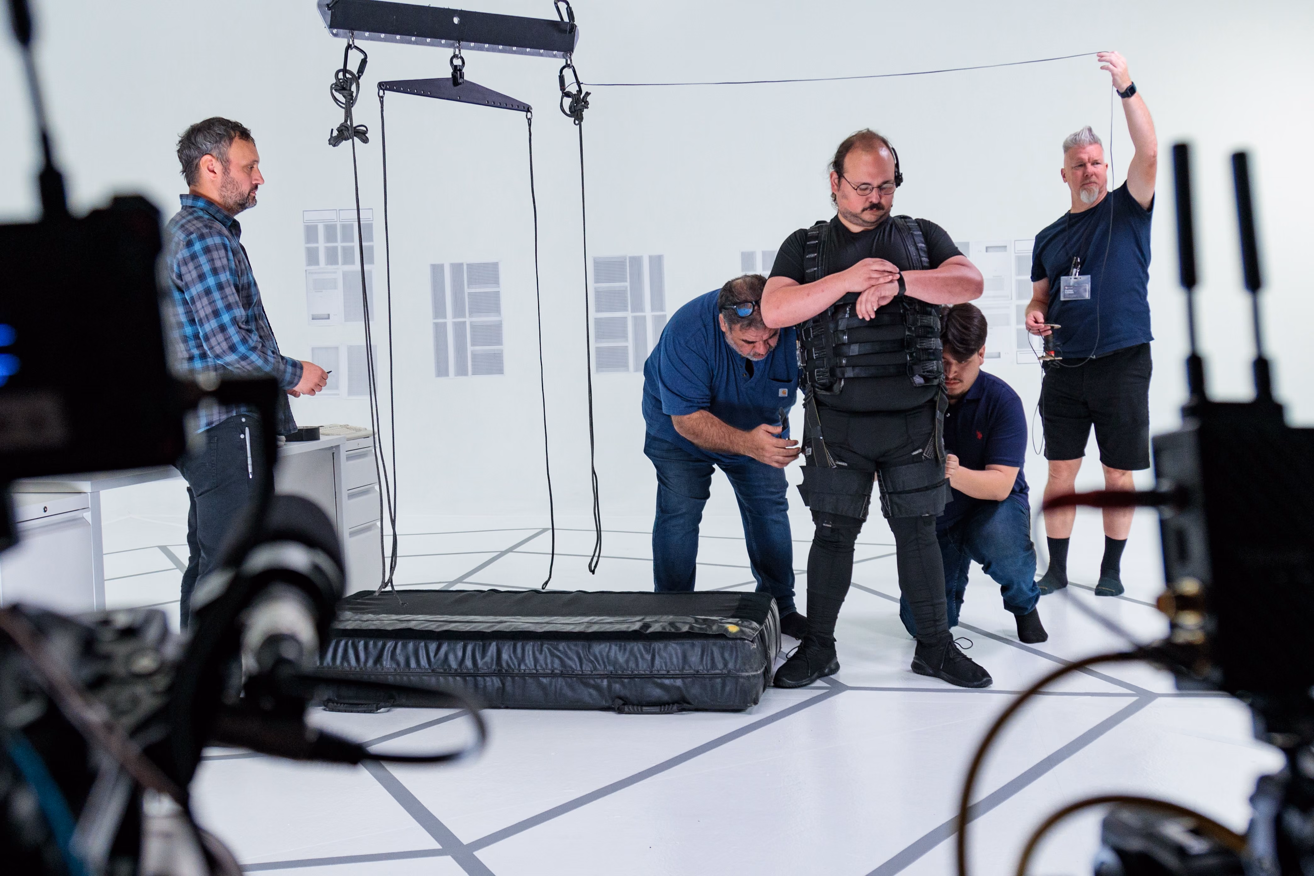 Crew fitting an actor into a stunt harness on a white studio stage