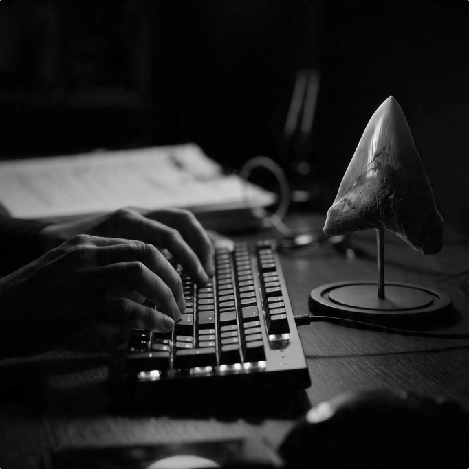 Black-and-white close-up of hands typing beside a mounted Megalodon tooth, representing discreet professional analysis and investigative work.