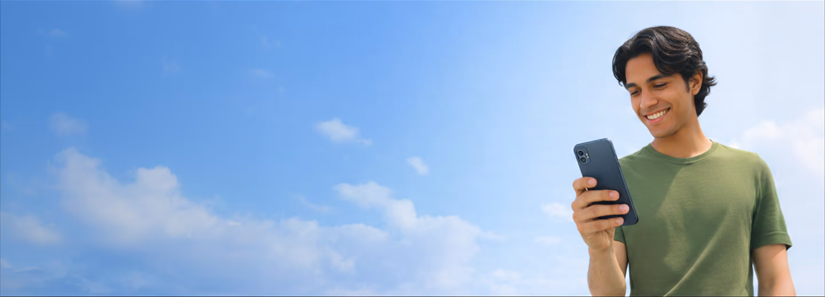 Joven sonriente vestido con camiseta verde mirando un teléfono móvil contra un cielo azul con nubes.