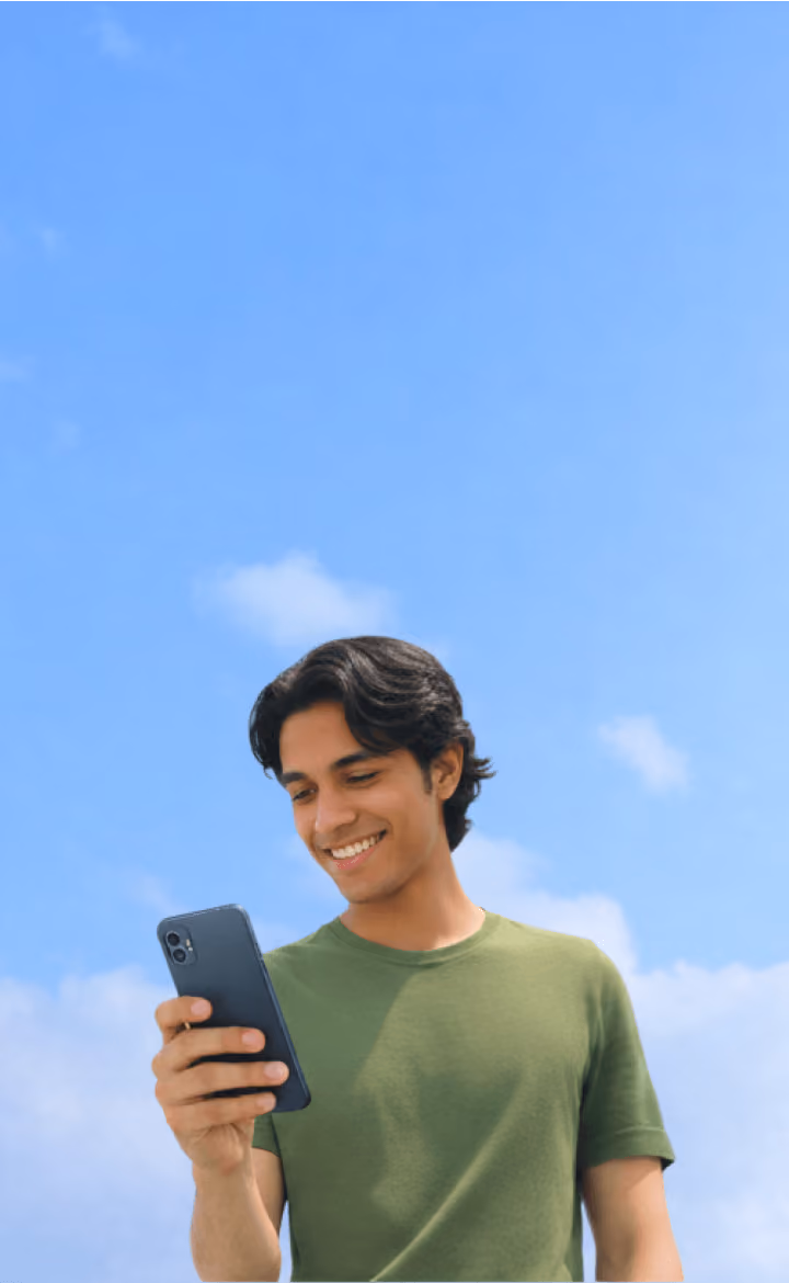Joven sonriente vestido con camiseta verde mirando un teléfono móvil contra un cielo azul con nubes.