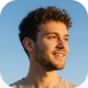 Joven sonriente con barba y cabello rizado mirando hacia la derecha con cielo azul de fondo.