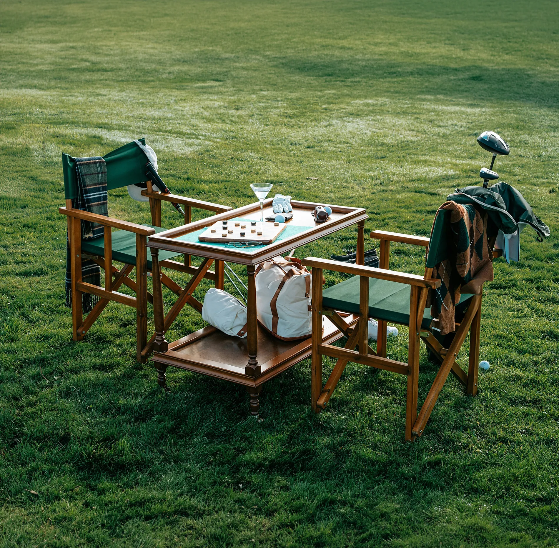 Two wooden chairs with green fabric and a table with checkers on grass with golf items nearby.