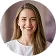 Smiling young woman with long light brown hair wearing a white top, standing against a purple background.