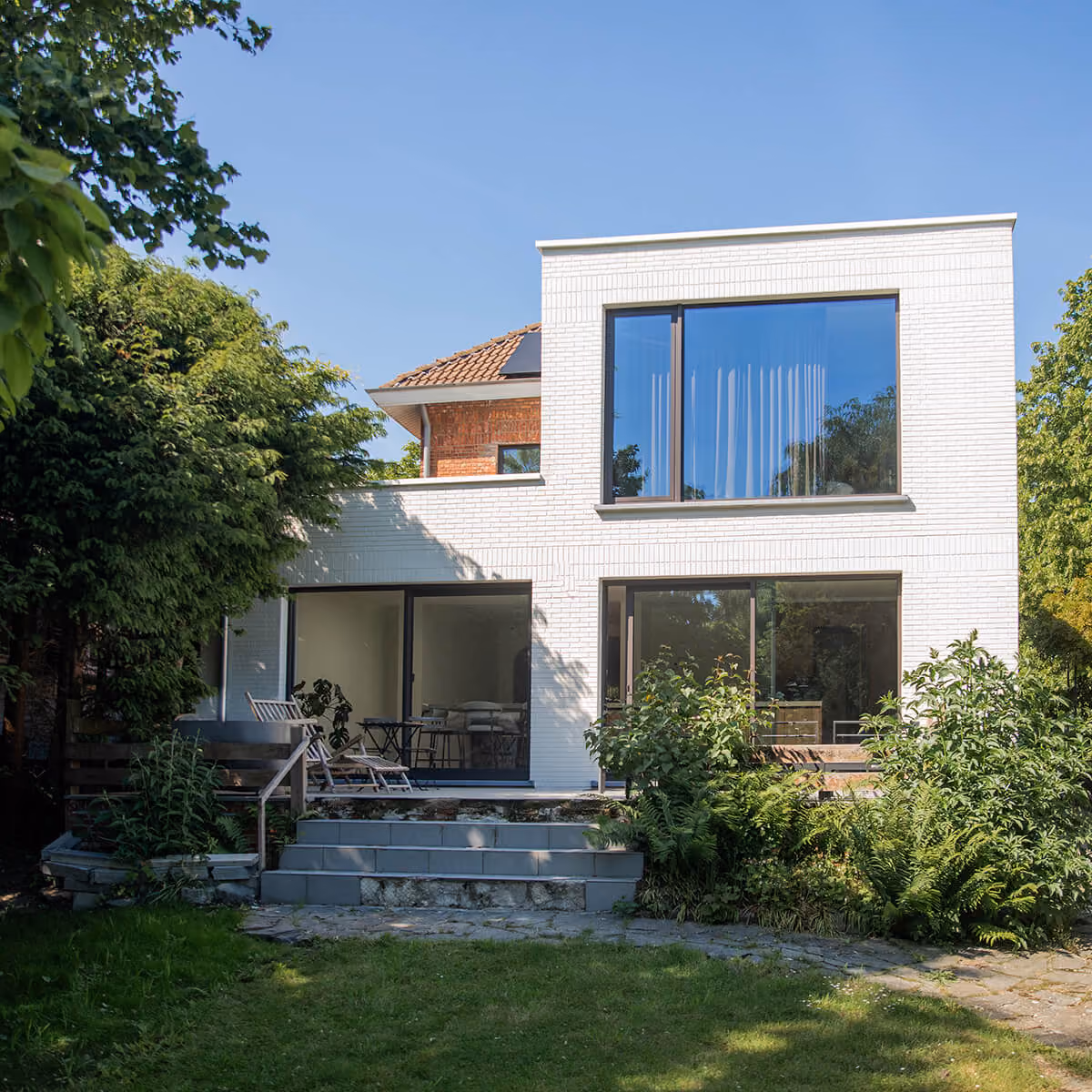 Achtergevel van een moderne witte woning met grote raampartijen en terras in een groene tuin