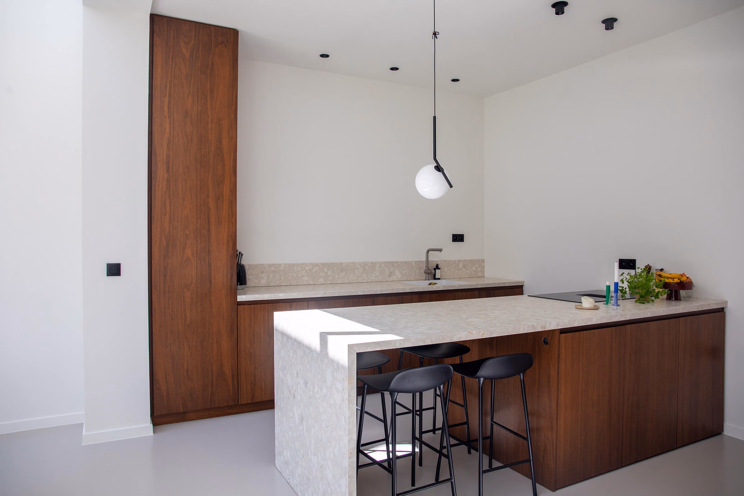 Moderne keuken met kastenwand in donker notenhout, een terrazzo-achtig werkblad in lichte tint, een kookeiland met zitgedeelte en drie zwarte barkrukken, een bolvormige hanglamp aan een zwart snoer en spotverlichting in het plafond
