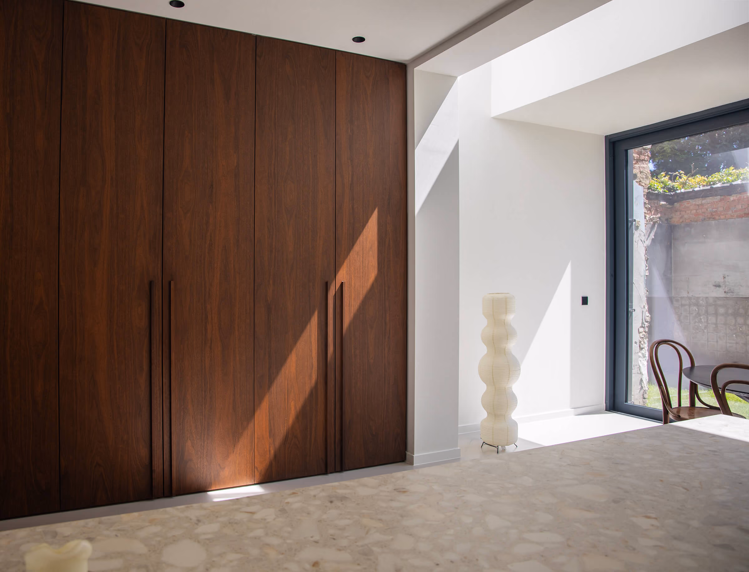Interieur met een hoge inbouwkast in notenhouten fineer met verticale greepsleuven, ernaast een lichte doorgang naar een ruimte met een golvende witte vloerlamp, terrazzovloer en zicht door een glazen deur op een tuin met stoelen.