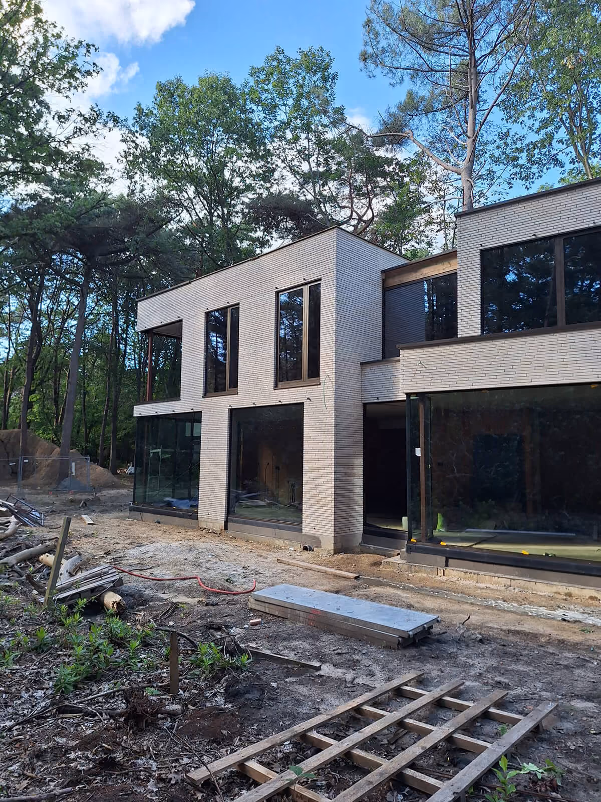 Moderne villa in aanbouw met witte baksteengevel en grote raampartijen, omringd door bos.