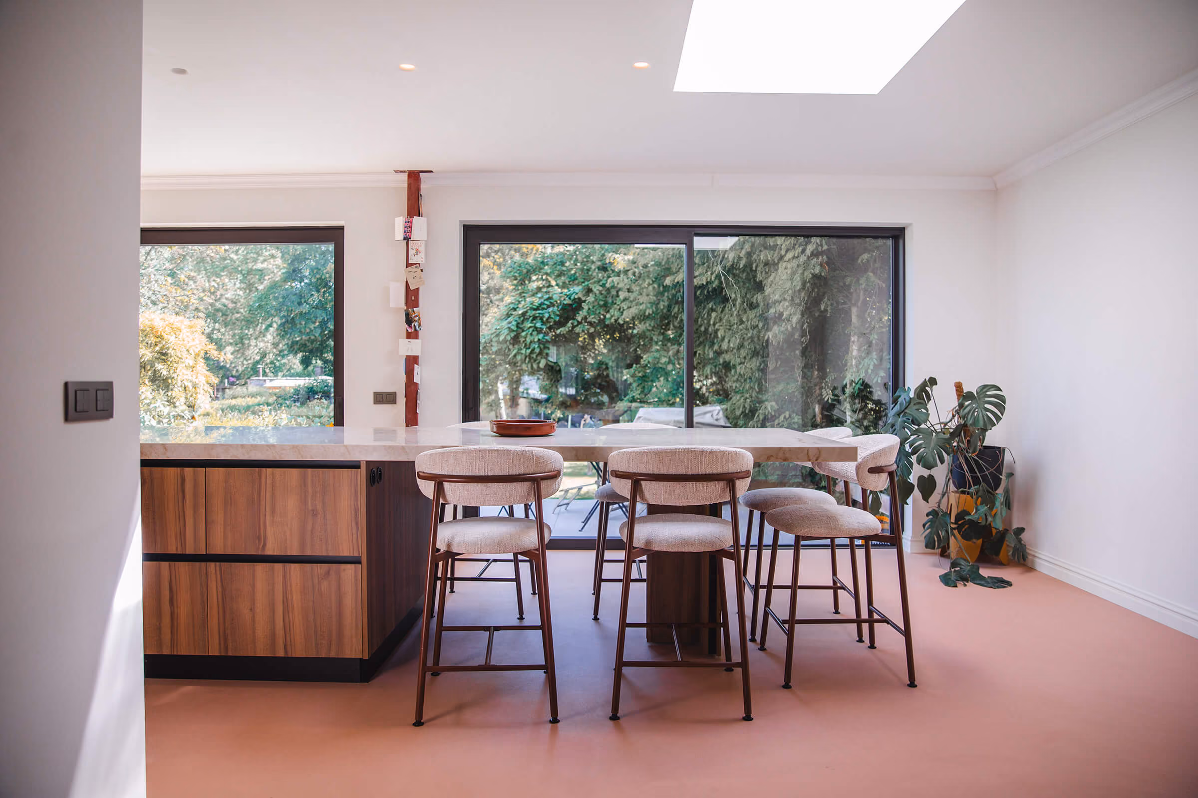 Open keuken met houten kastenwand, terrazzo eiland en barkrukken, roze vloer en grote schuifpui naar de tuin.