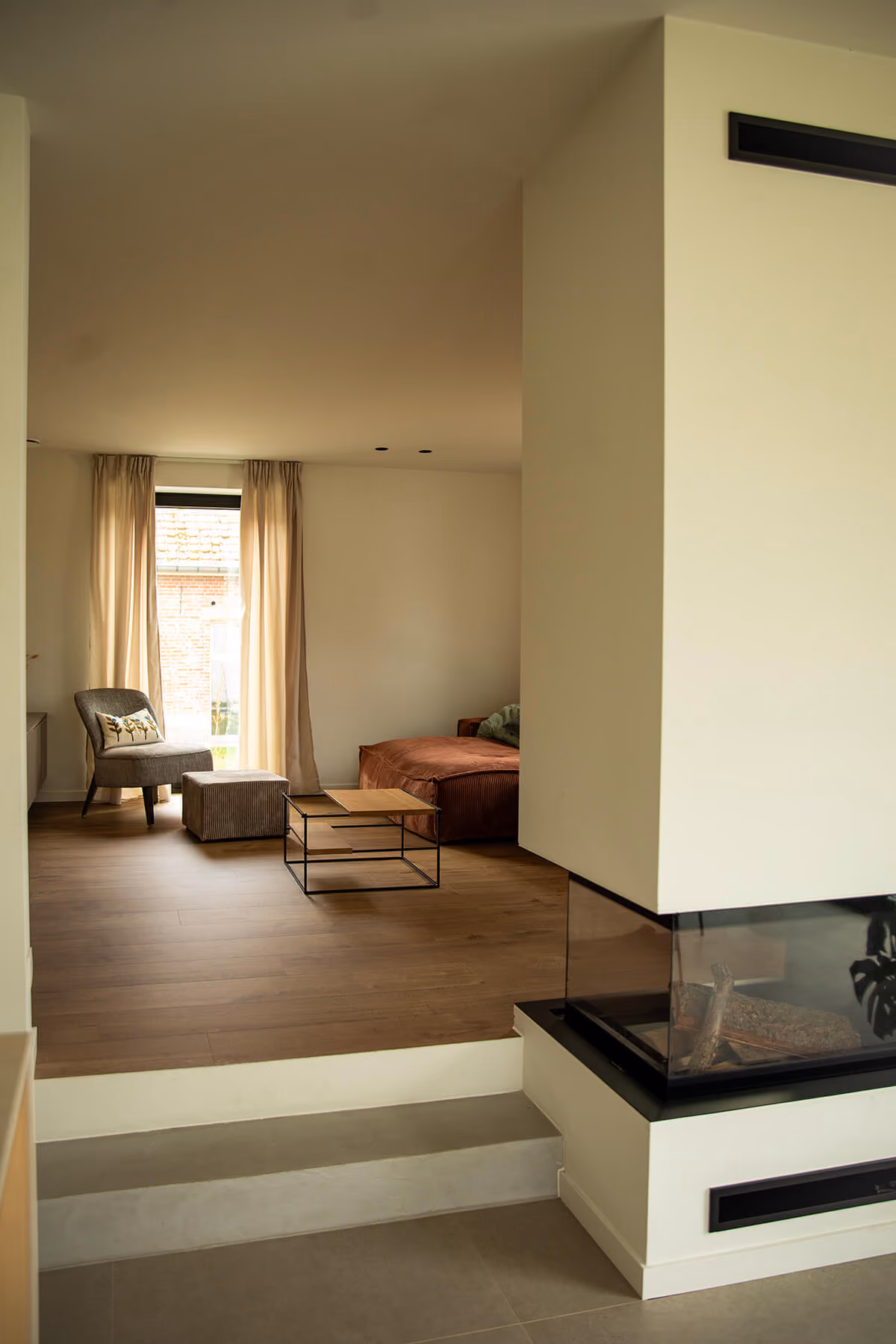 Woonkamer met houten vloer, zithoek en gashaard met glazen wand, verhoogd zitgedeelte.