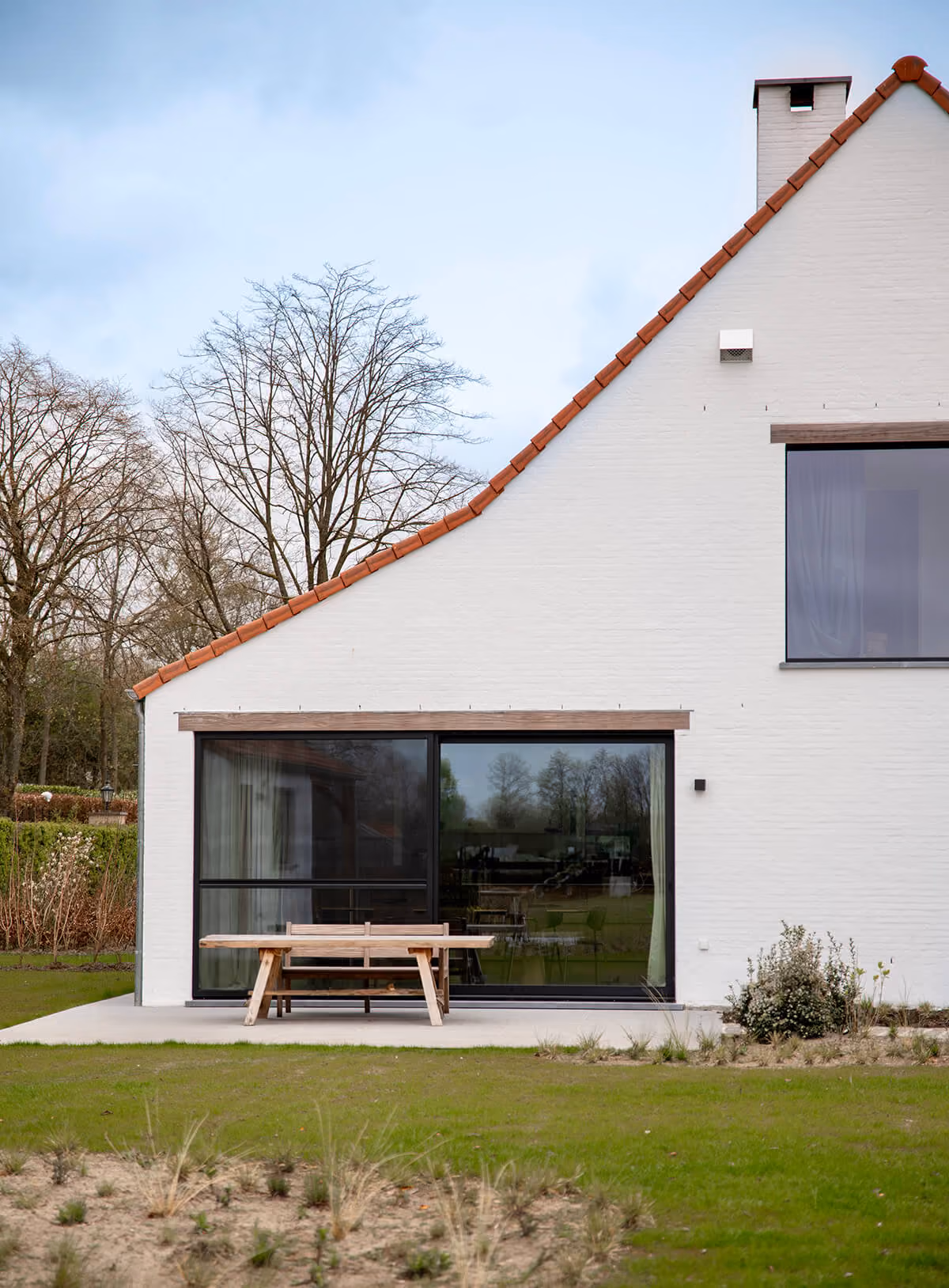Achterzijde van witte hoeve met puntgevel, rode dakpannen en grote glazen schuifpui naar de tuin.