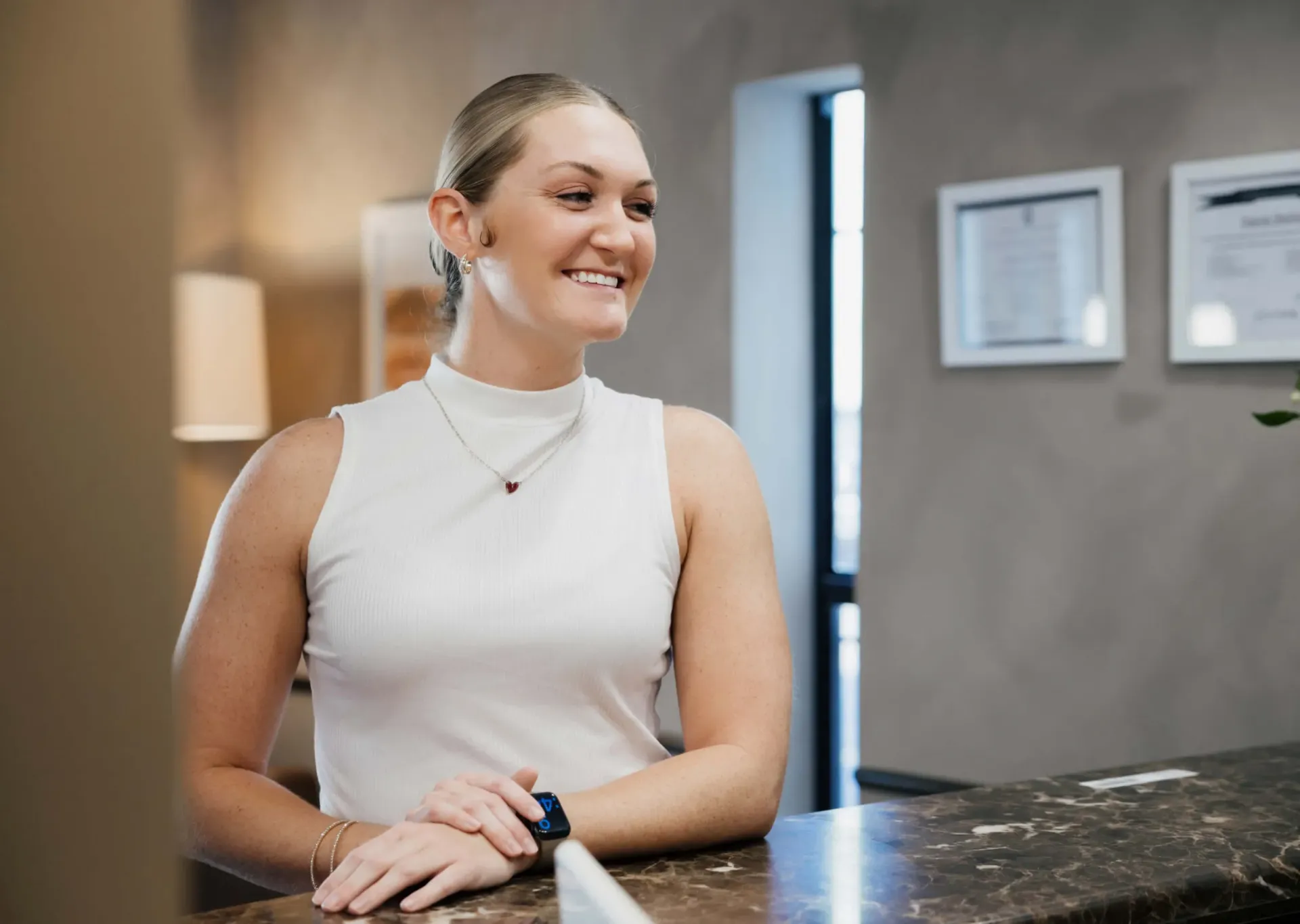 Smiling woman in white sleeveless top leaning on marble counter in a modern office setting.