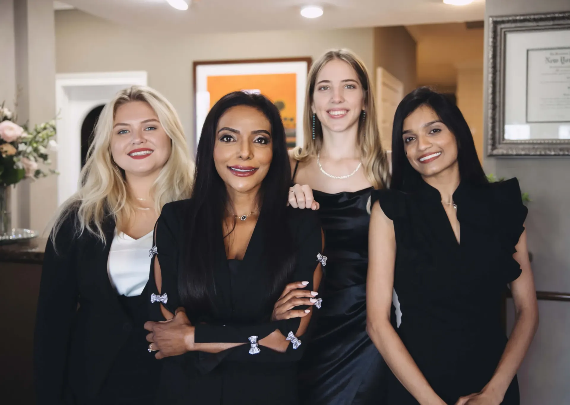 Four diverse professional women smiling, dressed in formal black and white attire indoors.