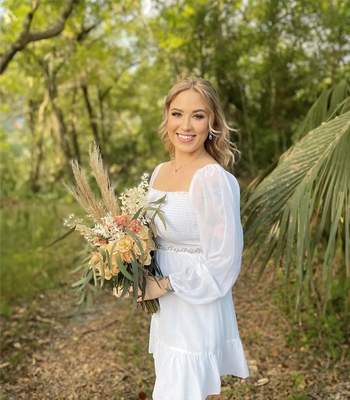 Smiling young woman in white dress holding a bouquet in a green forest setting.