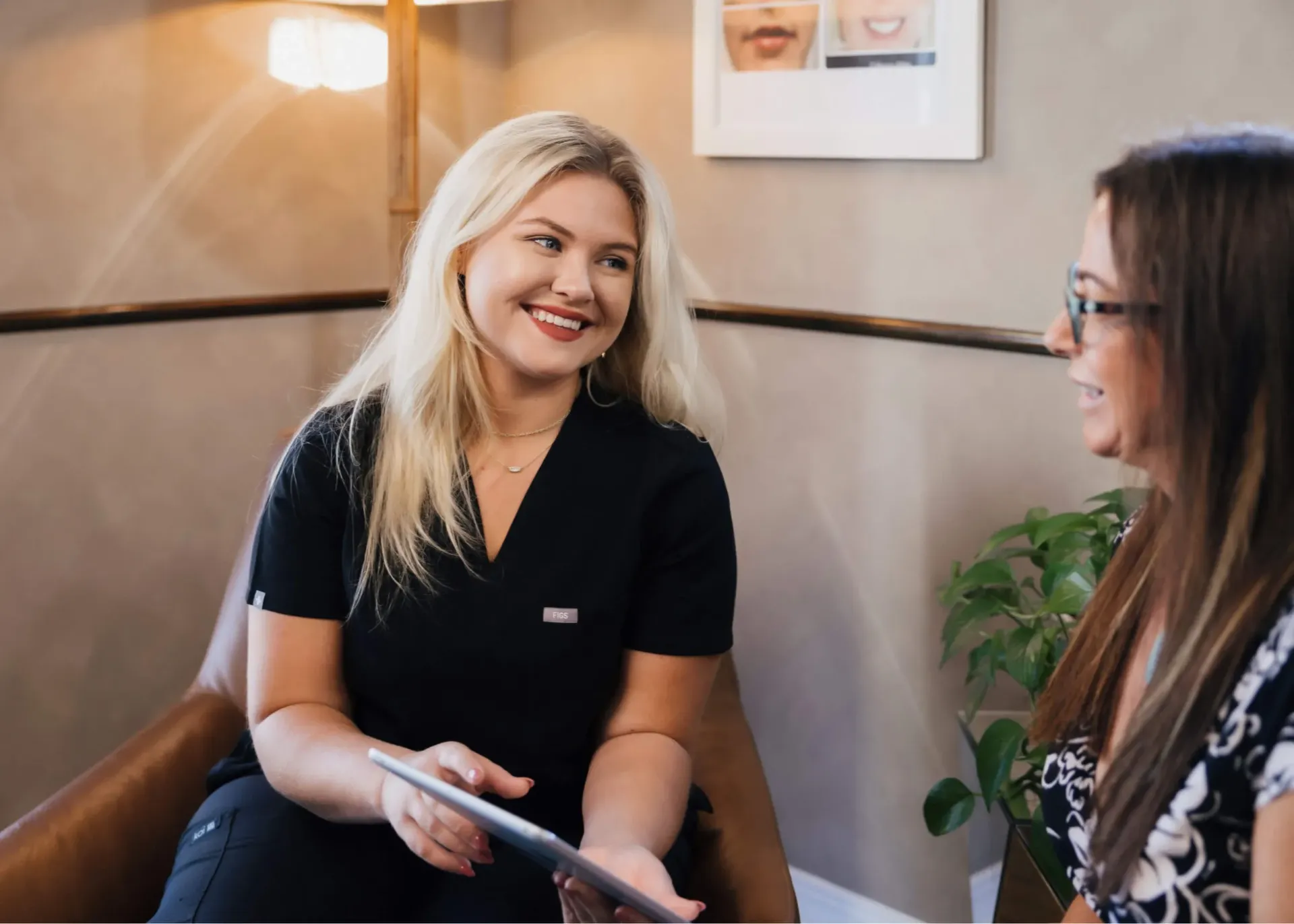 Blonde woman in black scrubs smiling while holding a tablet, talking to a woman with glasses.