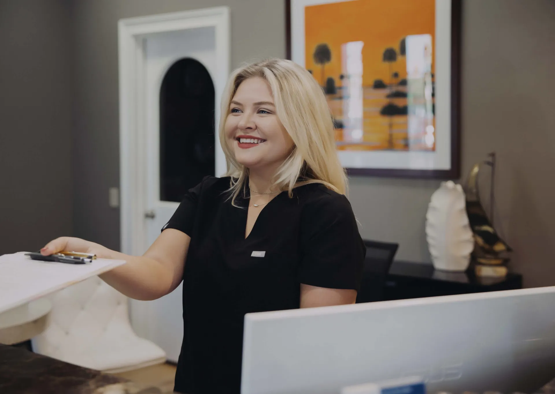 Smiling woman in black scrubs handing a clipboard at a reception desk in a modern office.