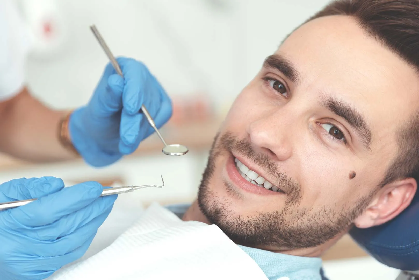 Smiling man reclined in dental chair with dentist's gloved hands holding dental tools nearby.