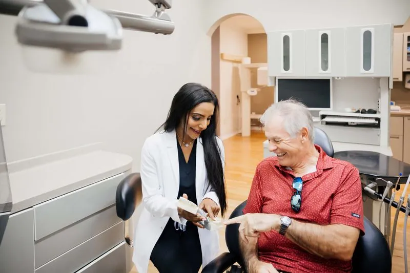 Dentist showing a dental model to an elderly man smiling in a bright dental office.