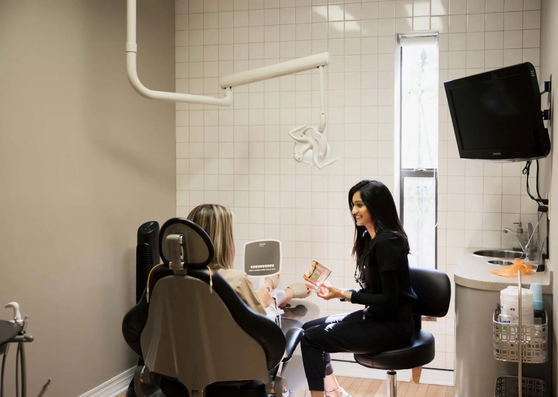 Dentist consulting a patient in a bright dental office with dental equipment and a TV on the wall.