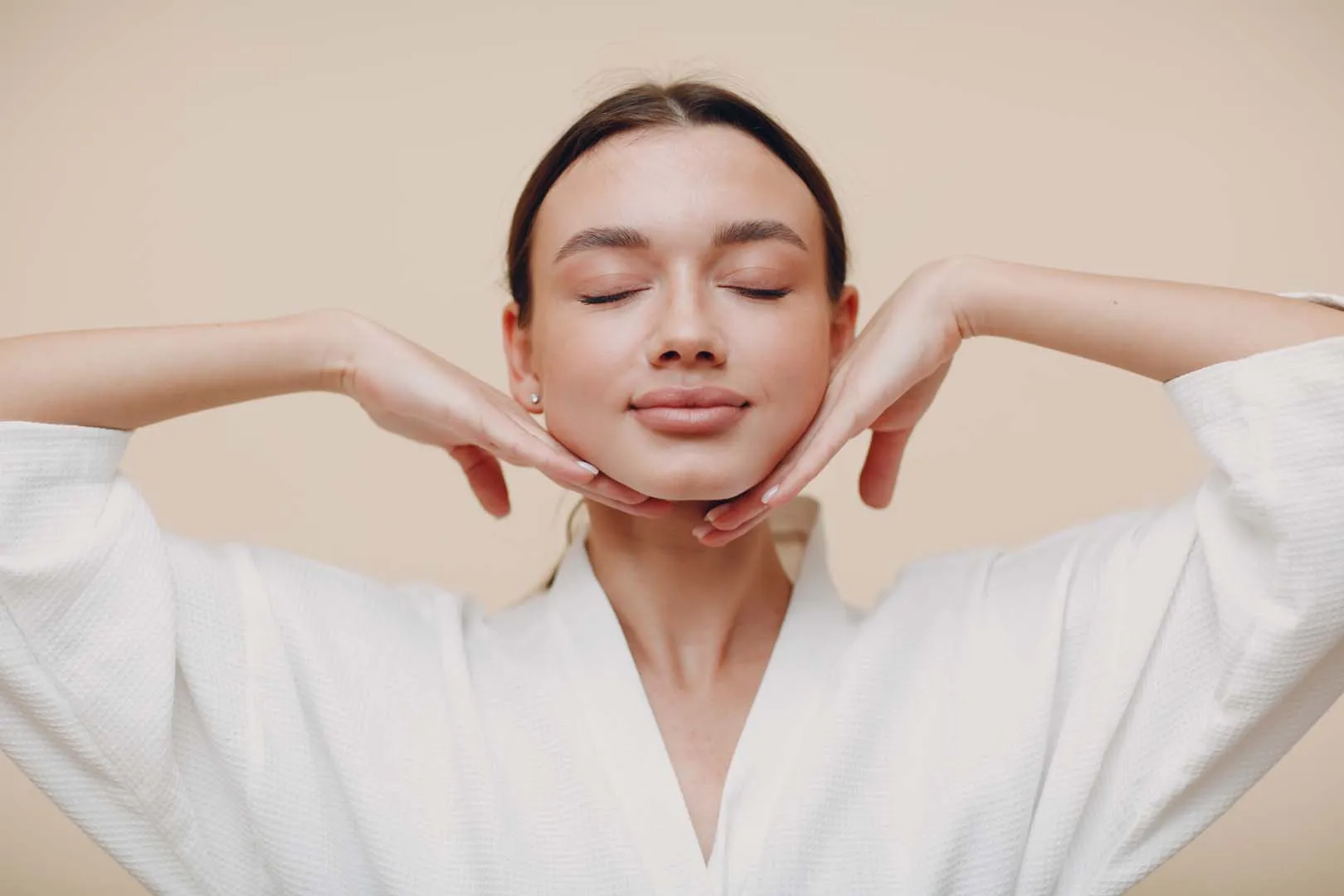 Young woman with closed eyes in a white robe poses with hands framing her face against beige wall.