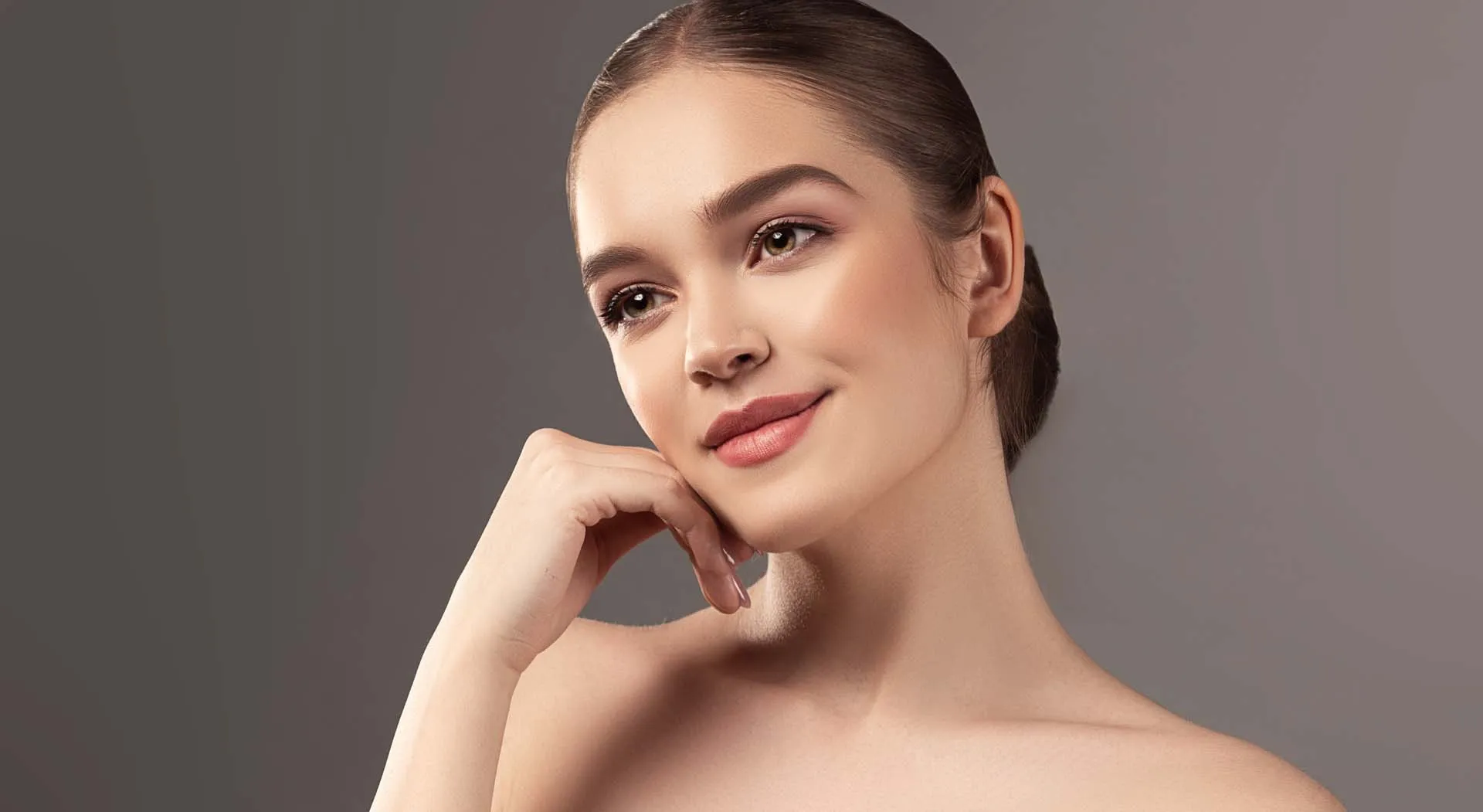 Young woman with smooth skin resting her chin on her hand against a plain gray background.